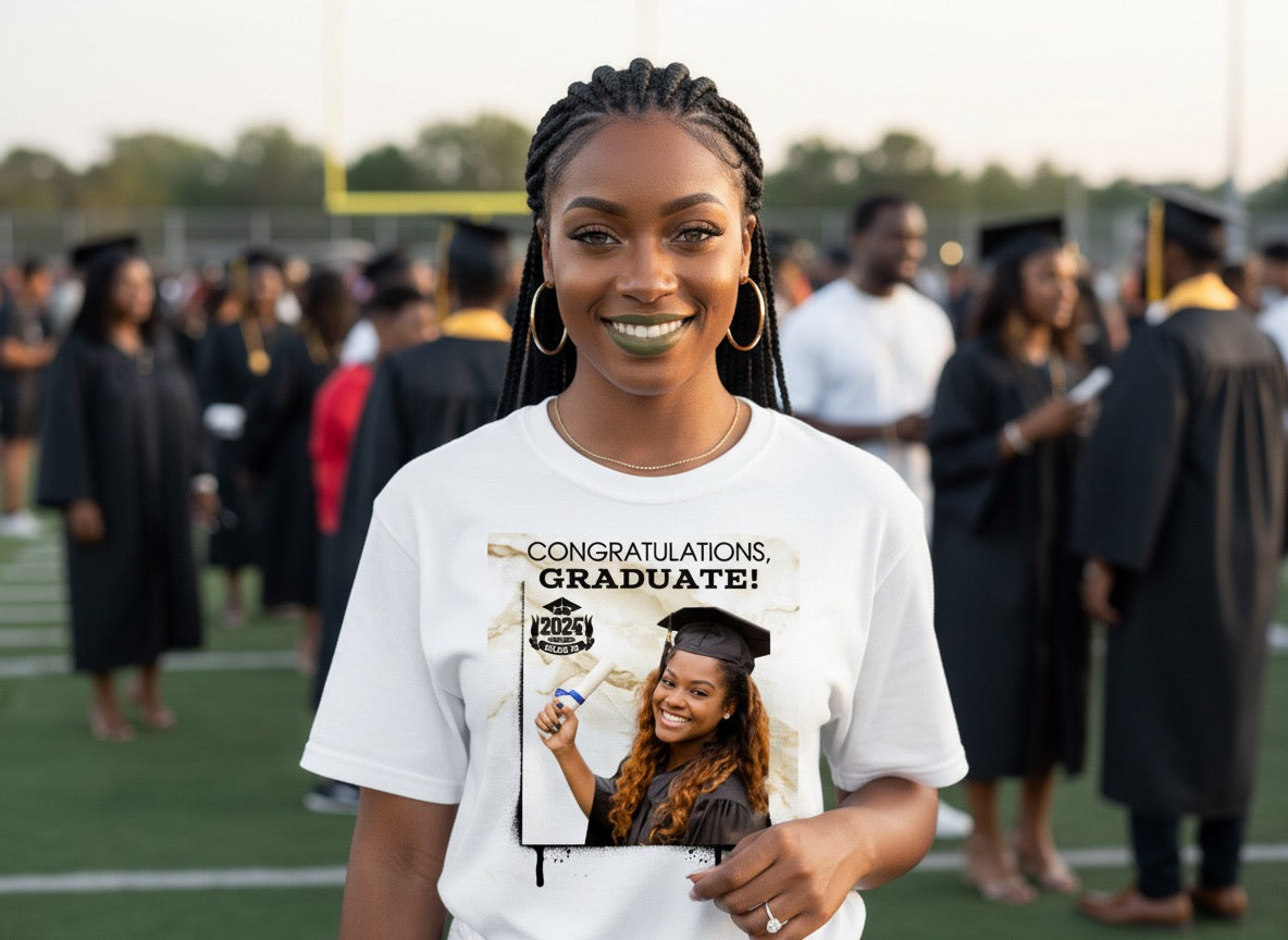 Woman wearing a white t-shirt with a graduation-themed graphic and text, standing on a sports field with graduation caps and gowns in the background.