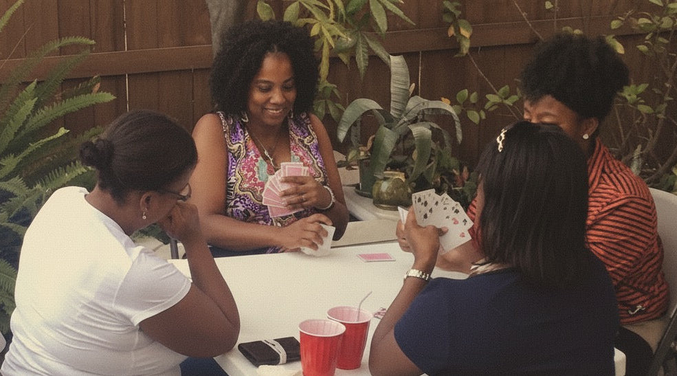Four women sitting around a table outdoors, playing cards and using a phone.