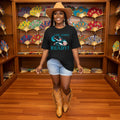 Woman in a black t-shirt with a colorful design, light blue shorts, and tan cowboy boots standing in front of a display of decorative fans.
