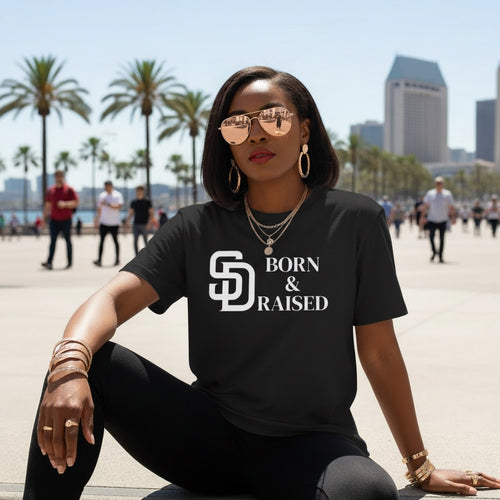 Woman wearing a black t-shirt with 'Born & Raised' text, sitting outdoors with palm trees and buildings in the background.