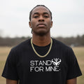 Man wearing a black t-shirt with 'Stand For Mine' text and a gold chain necklace outdoors.