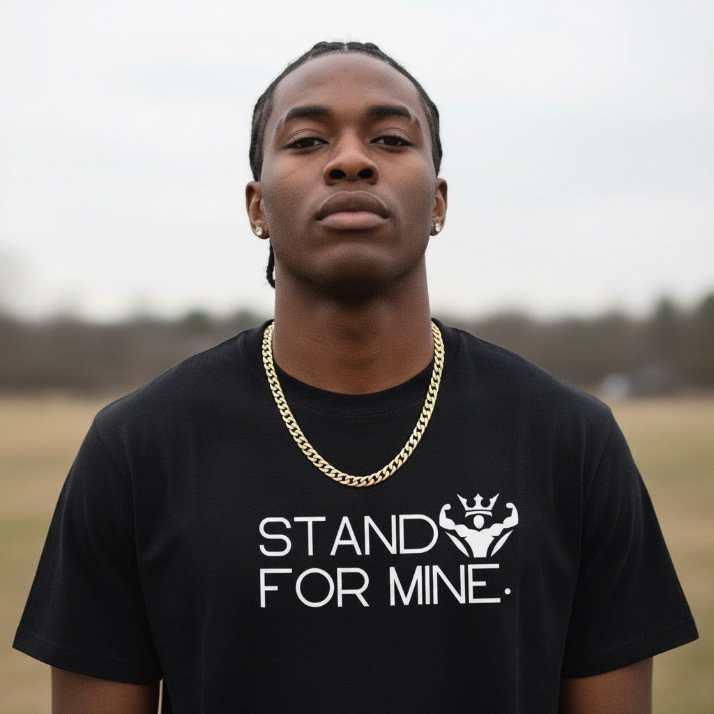 Man wearing a black t-shirt with 'Stand For Mine' text and a gold chain necklace outdoors.