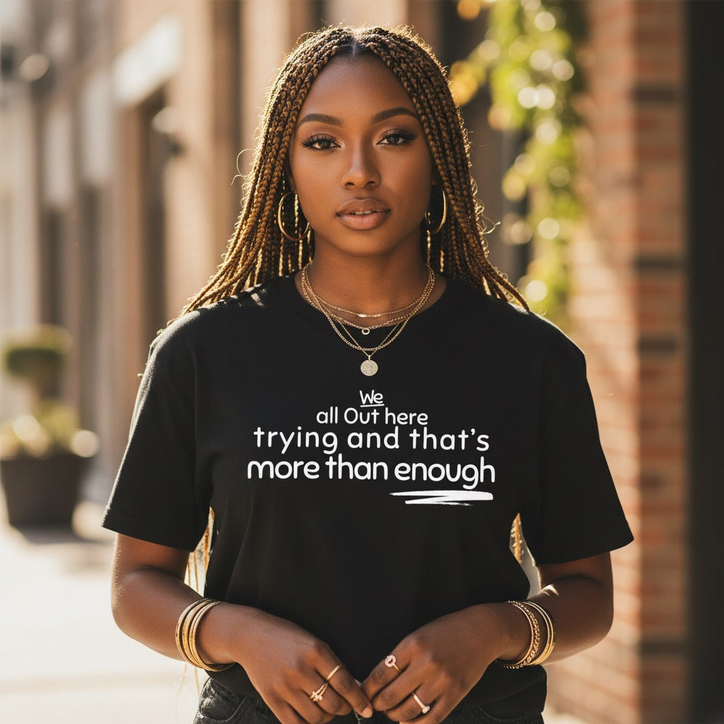 Black woman wearing the The Trying Is in the Triumph statement t-shirt featuring the phrase "We all out here trying and that's more than enough."