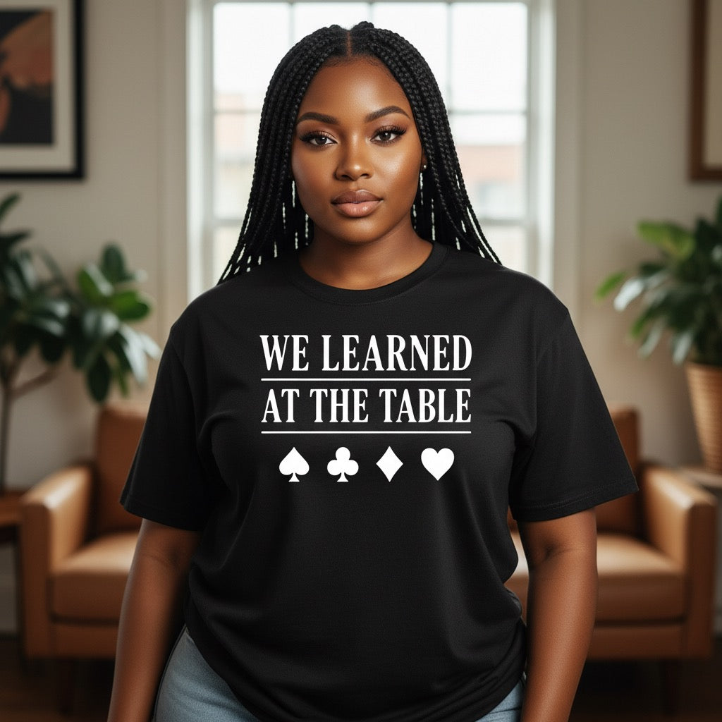 Woman wearing a black t-shirt with 'WE LEARNED AT THE TABLE' text and card symbols in a living room.