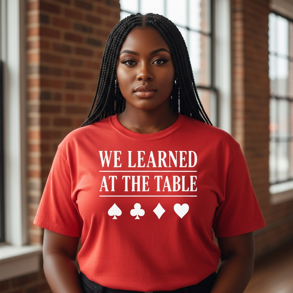 Person wearing a red t-shirt with 'WE LEARNED AT THE TABLE' text and symbols indoors.