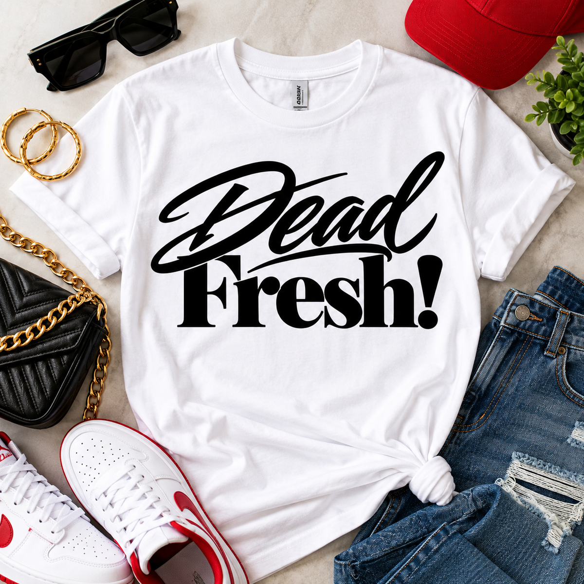 White Dead Fresh! graphic T-shirt with black lettering styled in a flat lay with sunglasses, gold jewelry, sneakers, and denim.